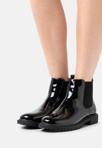 Bottes Chelsea en cuir verni noir avec panneaux latéraux élastiques et semelle en caoutchouc texturée, présentant une surface lisse et brillante ainsi qu'un bout arrondi.