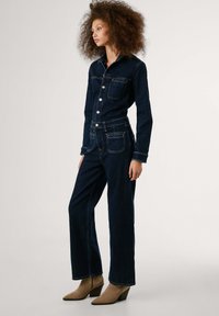 Mörk denim jumpsuit med en figursydd överdel, långa ärmar, krage, bröstficka och vida ben. Har kontrasterande sömmar och metalldetaljer.