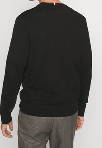 Pull en maille noir à manches longues, col rond et poignets côtelés. Le dos présente un logo tricolore près du col.