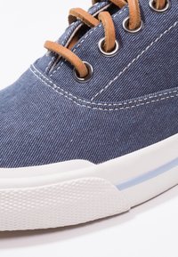 Denim-sneakers med en texturerad marinblå övre del, bruna lädersnören, silverhål och en vit gummisula med blå accent.