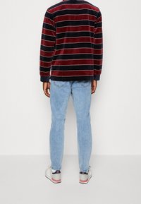 Man staat met zijn rug naar de kijker, gekleed in een rood-zwart gestreepte langemouwenshirt, lichtblauwe spijkerbroek en witte sneakers met roze accenten.