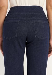 Takana näkymä henkilöstä, joka pitää yllään tummansinisiä farkkuja, joissa on kaksi takataskua, ja valkoista paitaa, joka on puettu sisään. Kädet rentoina sivuilla.