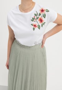 T-shirt bianco con ricami floreali colorati su un lato, abbinato a una gonna plissettata verde chiaro realizzata in tessuto morbido.