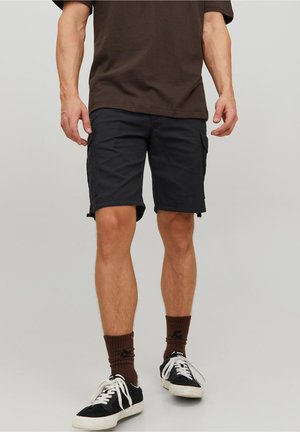 Homme portant un short cargo foncé, des chaussettes marron, des baskets noires et blanches, et un t-shirt marron foncé, debout sur un fond uni.