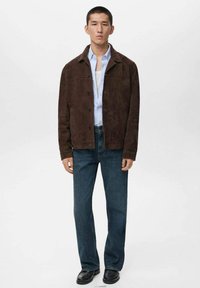 Giacca in suede marrone con colletto, chiusura a bottoni, indossata sopra una camicia azzurra chiara; abbinata a jeans in denim scuri e scarpe nere.