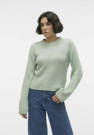 Lysegrønn ribbestrikket genser med rund hals og lange ermer, sammen med vide blå jeans. Stoffet ser mykt ut med en teksturert overflate.