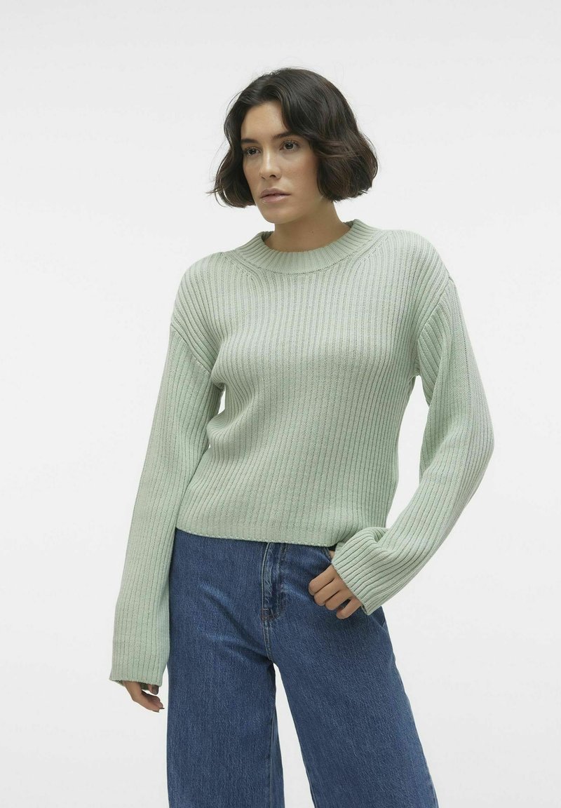 Pulover ribbed de culoare verde deschis, cu guler rotund și mâneci lungi, asortat cu blugi largi de culoare albastră. Țesătura pare moale, având un finisaj texturat.
