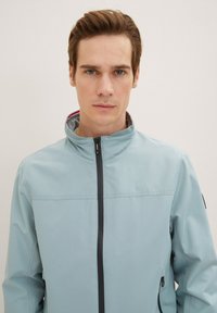 Giacca zip-up azzurro chiaro con colletto alto e zip nera. Presenta una texture liscia e un design discreto, adatta per un abbigliamento casual.