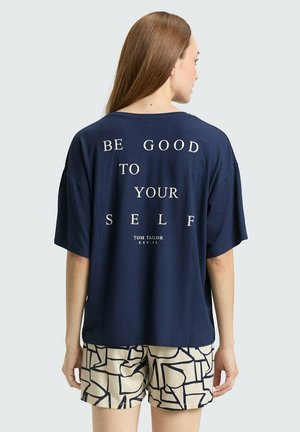 Kobieta z długimi brązowymi włosami nosi granatowy, oversize T-shirt z napisem "BE GOOD TO YOURSELF" i wzorzyste beżowe szorty, odwrócona tyłem.