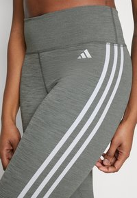 Leggings de sport gris avec une taille haute, présentant des rayures blanches sur les côtés et un petit logo Adidas sur la ceinture.