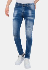Man in slim-fit blauwe versleten jeans met witte verfspatten en witte sneakers, staand met één hand in zijn zak.