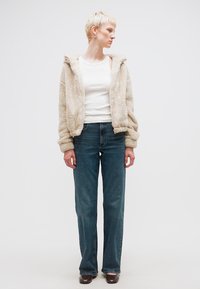 Άτομο με κοντά ξανθά μαλλιά που φοράει μπεζ φλις μπουφάν, λευκό τοπ, μπλε τζιν και καφέ παπούτσια, στέκεται μπροστά σε λευκό απλό φόντο.