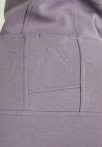 Sudadera con capucha morada que presenta una textura acanalada, costuras prominentes y un diseño minimalista. Detalle notable incluye un logo triangular bordado.
