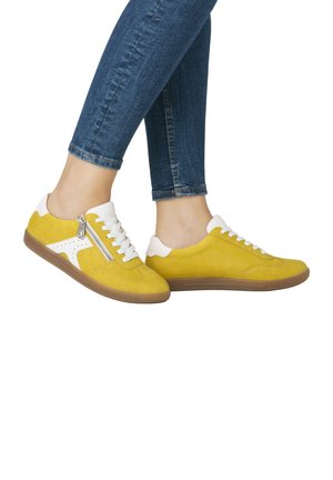 Gelbe Wildleder-Sneaker mit weißen Akzenten, ausgestattet mit einem seitlichen Reißverschluss und einer strukturierten braunen Gummi sole, seitlich zu sehen auf mit Jeans bekleideten Beinen.