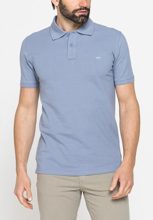 Uomo che indossa una polo azzurro chiaro a maniche corte e pantaloni beige, in piedi davanti a uno sfondo bianco uniforme.