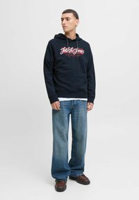 Granatowy hoodie z regulowanym sznurkiem i białe/czerwone logo, w połączeniu z luźnymi jasnoniebieskimi dżinsami i brązowymi masywnymi butami. Gładkie tło.