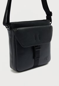 Μαύρη δερμάτινη τσάντα χιαστί με υφή, κλείσιμο με μαγνητικό καπάκι, ρυθμιζόμενο λουράκι και ανάγλυφο λογότυπο. Συμπαγής σχεδιασμός.