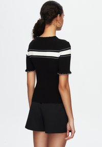 Top de punto negro con rayas blancas; mangas cortas con bordes rizados. Llevado con pantalones cortos negros; mostrado desde la parte trasera sobre un fondo blanco.
