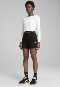 White long-sleeve crop top, black textured shorts with elastic waist, and black sneakers with white accents. Model stands against a gray background.