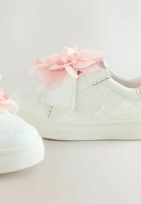 Valkoiset lenkkarit, joissa on teksturoitu pinta ja vaaleanpunainen kerroksellinen kukka aksentti sivussa. Varustettu säädettävillä hihnoilla ja tasaisella kumipohjalla.