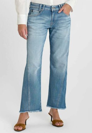 Vrouw draagt lichtblauwe rechte jeans met onafgewerkte zoom, bruine sandaaltjes met hak en open tenen, en een wit, langärmelig overhemd dat is ingestopt.