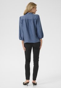 Blaue Chambray-Bluse mit gerafftem Rücken und Rüschkragen, kombiniert mit schwarzen Skinny Jeans und schwarzen Absatzmules.