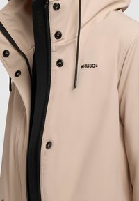 Veste softshell beige avec fermeture éclair, quincaillerie noire, capuche ajustable et logo brodé. Texture lisse et coupe ajustée.