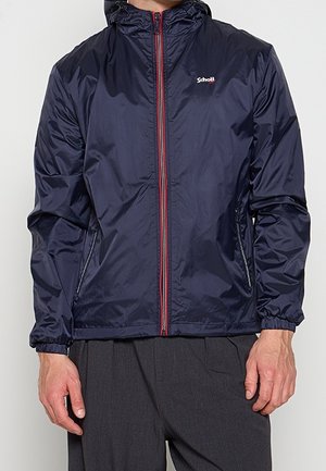 Homme portant une veste à capuche bleu marine avec une bordure de fermeture éclair rouge et le logo Schott blanc sur la poitrine, associée à un pantalon gris foncé.