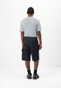 Mørke denim cargo shorts med flere lommer, gule sømmer og en avslappet passform. Kombinert med en lys grå t-skjorte og svarte sokker.