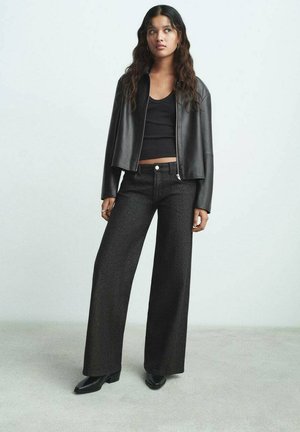 Junge Frau mit langen Haaren, die eine schwarze Lederjacke, ein schwarzes Croptop, weit geschnittene schwarze Hosen und schwarze Stiefeletten vor einem schlichten Hintergrund trägt.