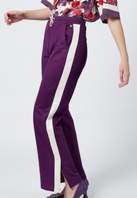 Fioletowe spodnie dresowe z białymi bocznymi paskami, z elastyczną talią ze sznurkiem, kieszeniami po bokach i rozcięciami na dole. Miękka tekstura materiału.