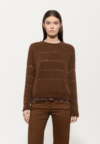 Brauner Strickpullover mit horizontalen Streifen und strukturiertem Muster. Über einem gemusterten Oberteil getragen, kombiniert mit passenden braunen Hosen.