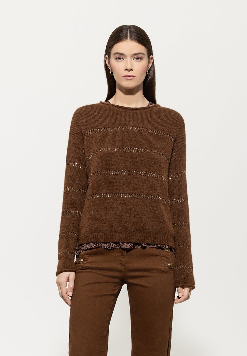 Brauner Strickpullover mit horizontalen Streifen und strukturiertem Muster. Über einem gemusterten Oberteil getragen, kombiniert mit passenden braunen Hosen.