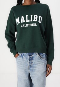 Zelená mikina s bílým potiskem "MALIBU CALIFORNIA", která má kulatý výstřih a volné rukávy, kombinovaná se světle modrými džínami.