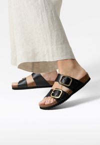 Zwarte tweeband-sandalen met grote gouden gespen, kurken voetbed en gestructureerde zwarte loopzool. Gemaakt met een lichtbeige linnen rok.