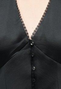 Μαύρο ένδυμα με V-λαιμόκοψη διακοσμημένη με μαύρη δαντέλα, που έχει μια σειρά από μαύρα κουμπιά κατά μήκος του κεντρικού μπροστινού μέρους.