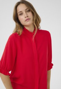 Blusa rossa con un colletto alto arricciato e maniche corte a sbuffo. Realizzata in tessuto morbido con una vestibilità ampia e dettagli con bottoni sul davanti.