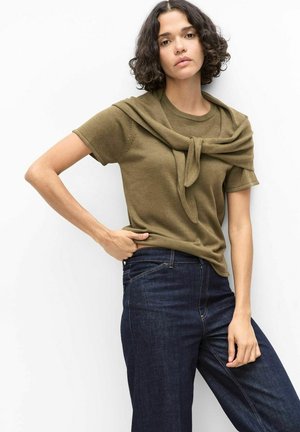 Vrouw met kort krullend haar, gekleed in een olijfgroene top met korte mouwen en donkerblauwe jeans, met een bijpassende trui om haar schouders geknoopt.