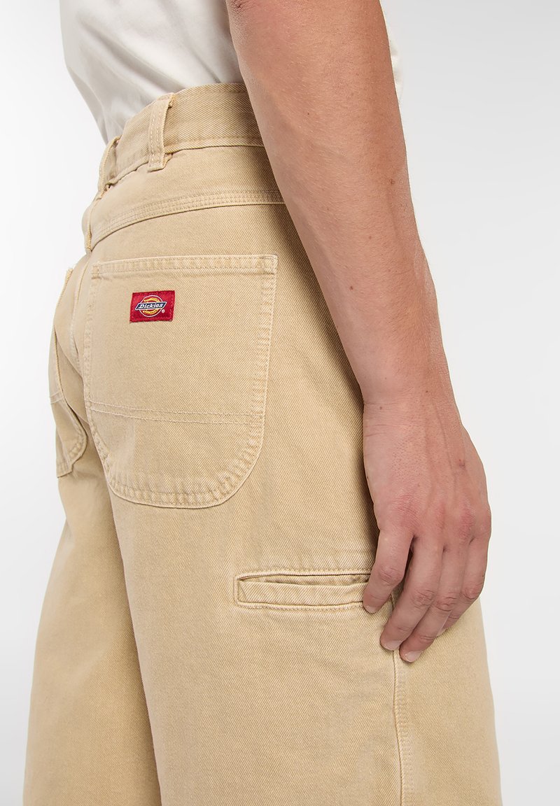 Pantalones de trabajo beige con un corte holgado, que presentan un bolsillo parche y una tela con textura sutil. Etiqueta roja cerca del bolsillo trasero.