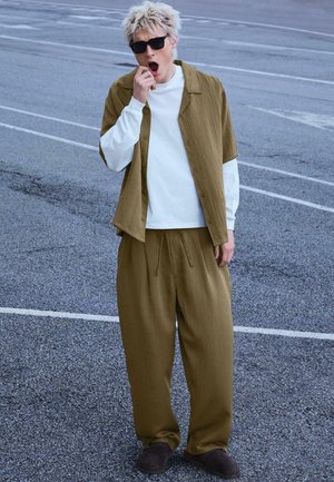 Mladi človek razmršene blond lase in sončna očala zeha, nosi ohlapne rjave hlače in kratko rjavo majico preko bele majice z dolgimi rokavi.