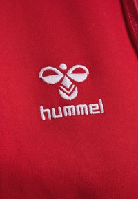 Rode stof heeft een witte geborduurde logo met een gestileerd bijontwerp boven het woord "hummel" in een vetgedrukt, kleine letters font.