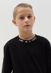 Zwarte sweatshirt met ronde hals, met daarop de witte tekst "wake up to reality" op de kraag en vlamontwerpen op de mouwen.