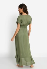 Μακρύ φόρεμα σε ελιά πράσινο χρώμα με ομαλή, ρέουσα υφή, κοντά μανίκια, άνετη εφαρμογή, σούρα στη μέση και φούστα που ανοίγει προς τα κάτω.