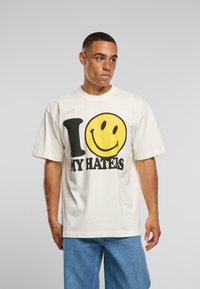 Ungdomsmann med kort hår iført en hvit T-skjorte med et stort gult smilefjes og teksten "I [smiley] MY HATERS", kombinert med blå jeans.