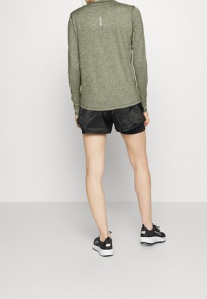Personne portant une chemise olive à manches longues, un short noir superposé et des baskets Nike noires, debout face à un mur blanc uni.