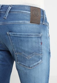 Ljusblå denimjeans med strukturerad yta. Har en brun läderlapp vid midjan och en detaljerad design på bakfickan.