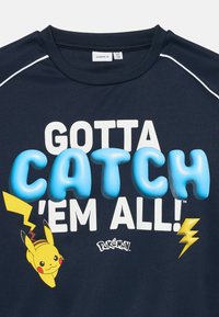 Námornícky modrá košeľa s textom "MUSÍM ICH VŠETKY ZACHYTÍM!", 3D modrým "CATCH", Pikachu s bleskami a logom Pokémon na prednej strane.