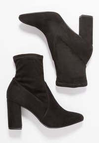 Botas de tobillo de ante negro con punta afilada y tacón cuadrado. Presentan un caño caído con acentos de costura diagonal.