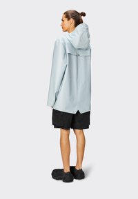 Chaqueta impermeable de color azul claro con capucha, corte relajado, ventilación trasera y dobladillo asimétrico, combinada con pantalones cortos negros y zapatos negros texturizados.