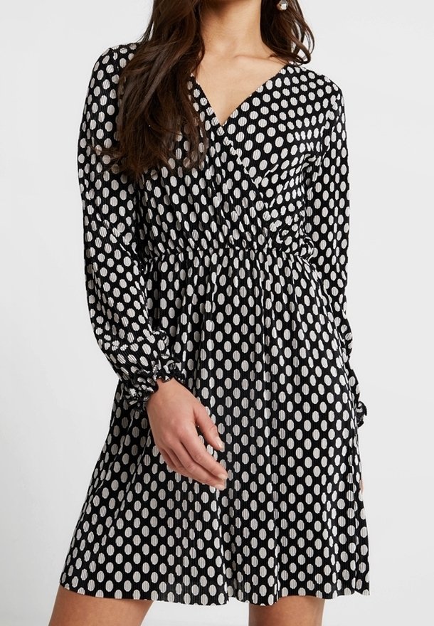 Schwarzes Kleid mit langen Ärmeln, einem V-Ausschnitt, elastischem Bund und weißen Punkten. Der Stoff wirkt glatt mit einem subtilen Glanz.